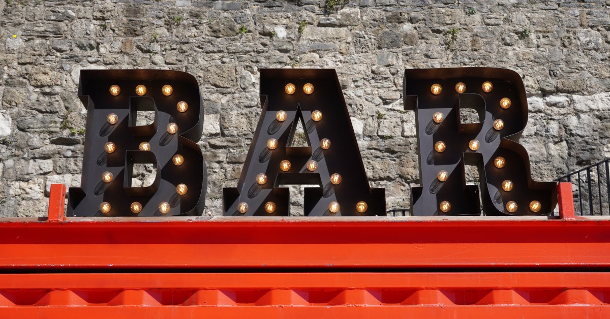 A black retro sign that says "bar" in bold letters sits on top of a red platform in front of a gray stone wall.