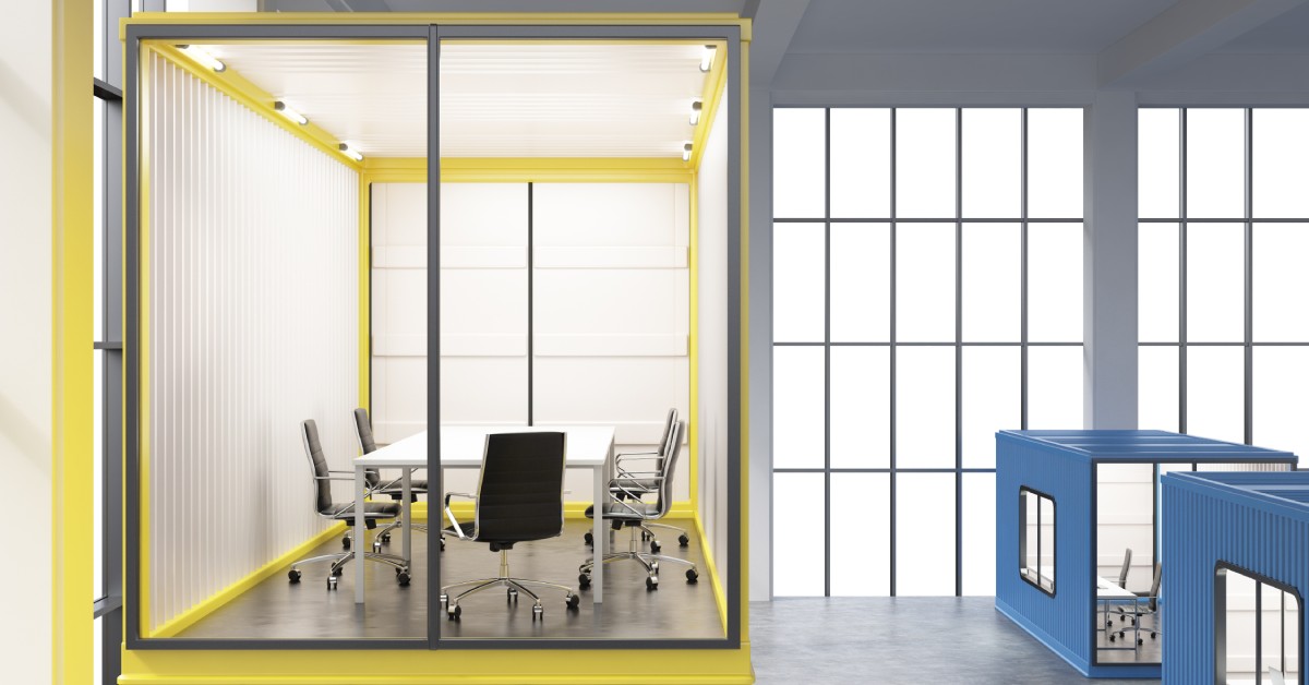 A conference room made out of a yellow shipping container sits in a large building. Individual container offices sit nearby.