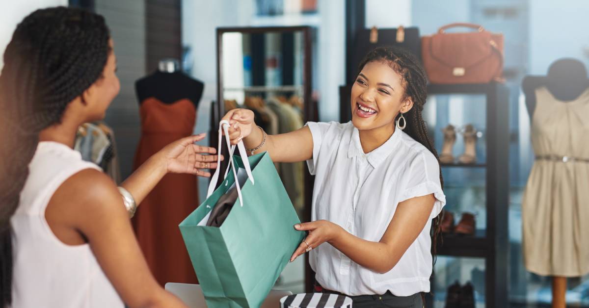 A store owner smiles as she hands a shopping bag to a customer over the counter. Clothing merchandise is in the background.