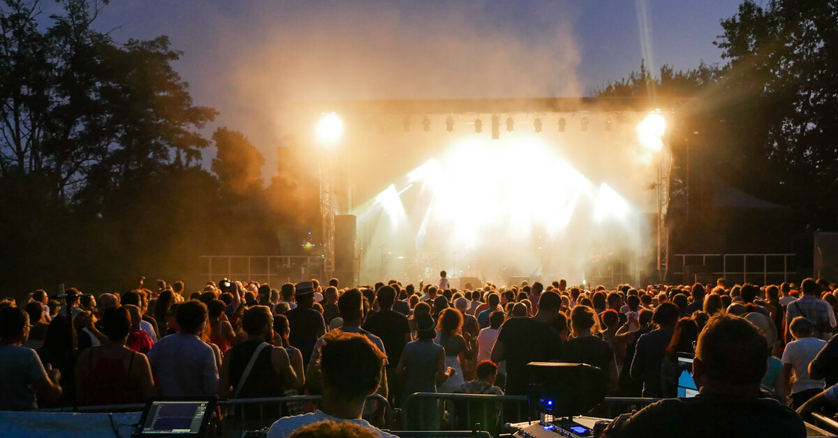 A large crowd faces a brightly-lit stage during an outdoor concert at night. The stage is bordered by trees.