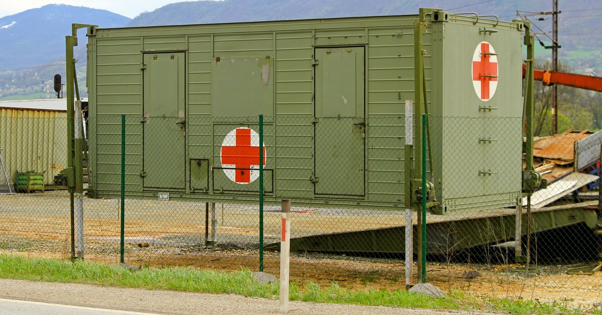 A green shipping container with a red cross logo sits alongside fencing with mountains and housing construction behind it.
