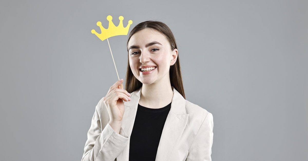 A woman with brown hair wearing a beige blazer smiles and holds up a photo booth prop of a yellow crown.